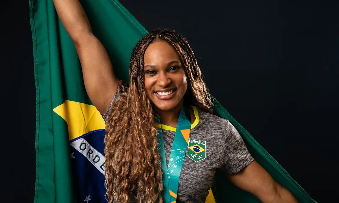 ginasta Rebeca Andrade com uniforme do Brasil, medalha pendurada no pescoço e bandeira do Brasil nas costas, sorrindo para a câmera, de cabelo solto