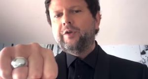 Selton Mello de roupa preta, falando para a câmera e mostrando anel