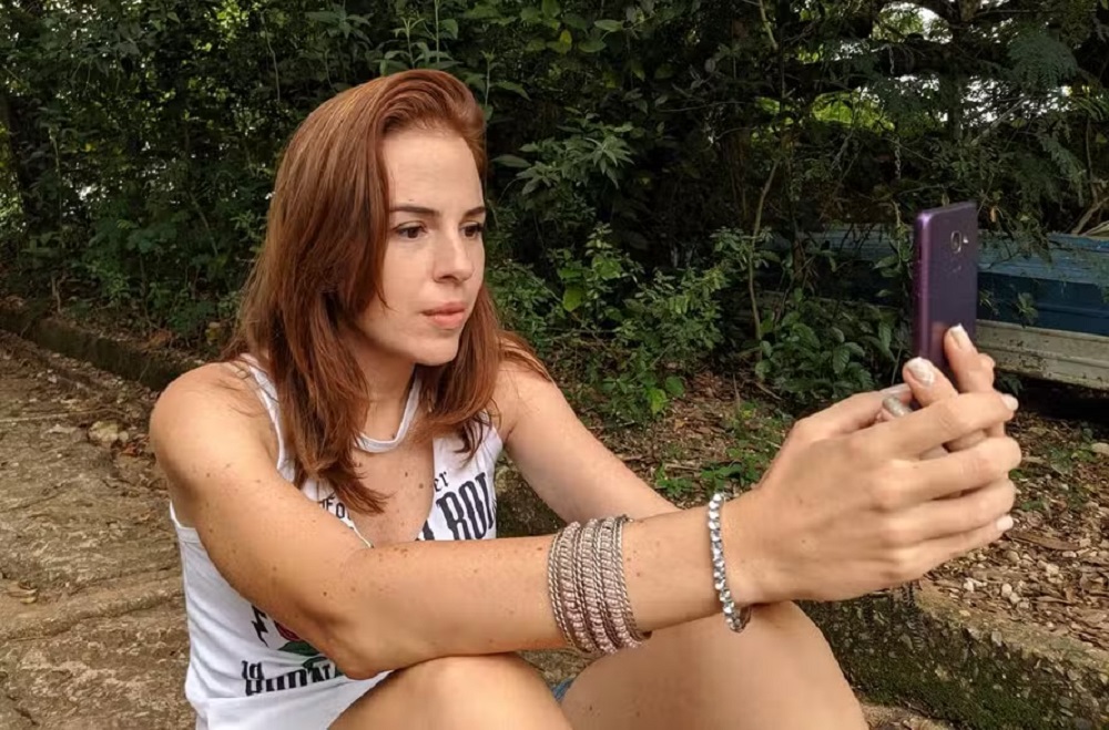 Suzane von Richthofen séria, de cabelo solto e roupa branca sem mangas, mexendo no celular sem olhar para a câmera