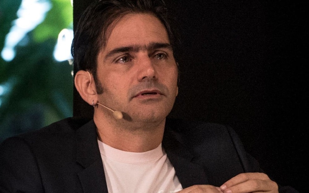 Vinicius Coimbra falando sem olhar para a câmera, sério, de roupa branca e preta