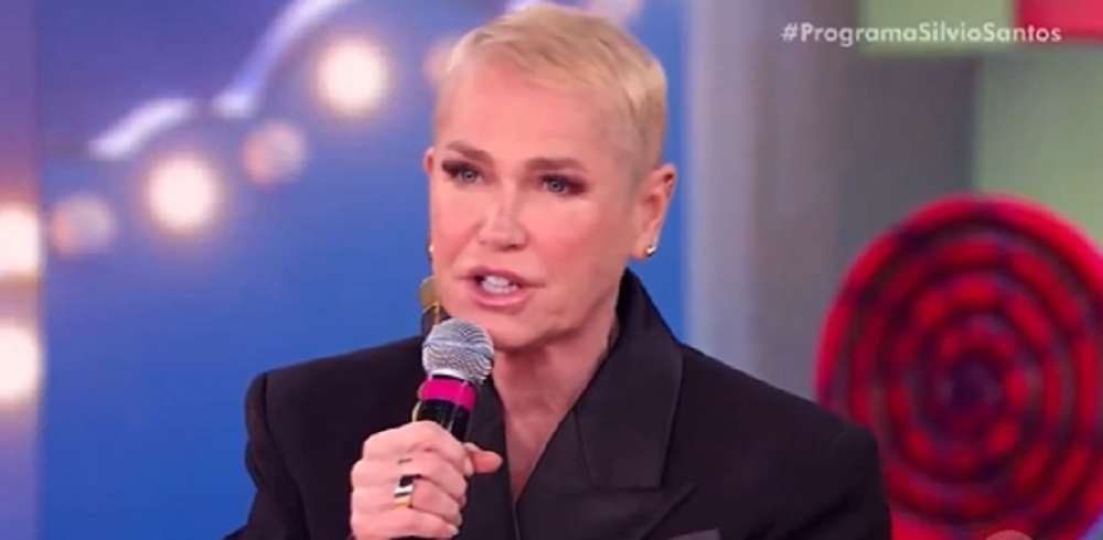 Xuxa Meneghel em participação no Programa Silvio Santos com Patrícia Abravanel, de roupa preta, falando em microfone, sem olhar para a câmera, sério