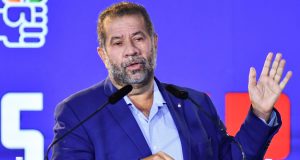 Carlos Lupi de roupa social azul, sério, falando em microfone e gesticulando, sem olhar para a câmera