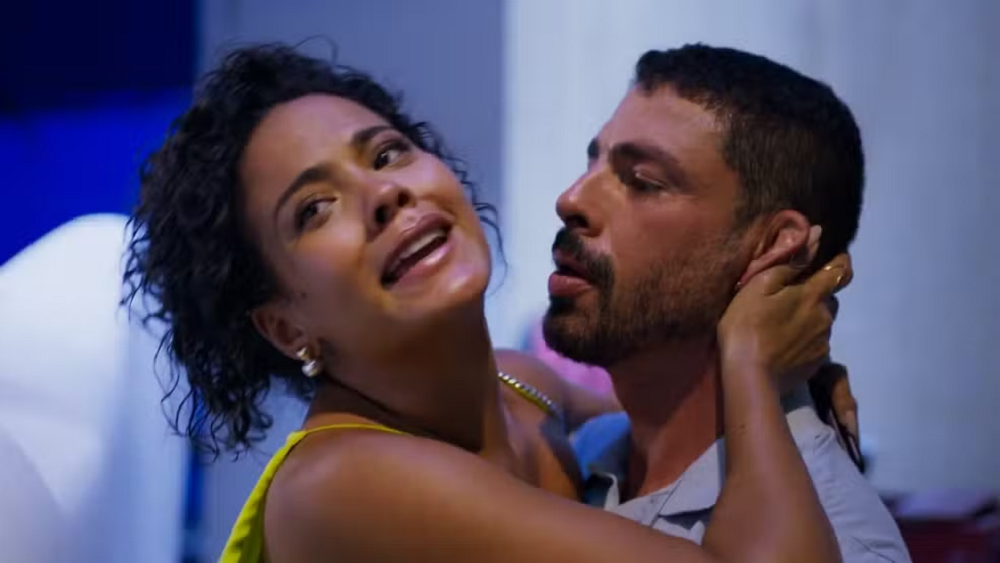 Os atores Bella Campos e Cauã Reymond abraçados em cena do remake de Vale Tudo, ele sério de perfil e ela com expressão de desespero