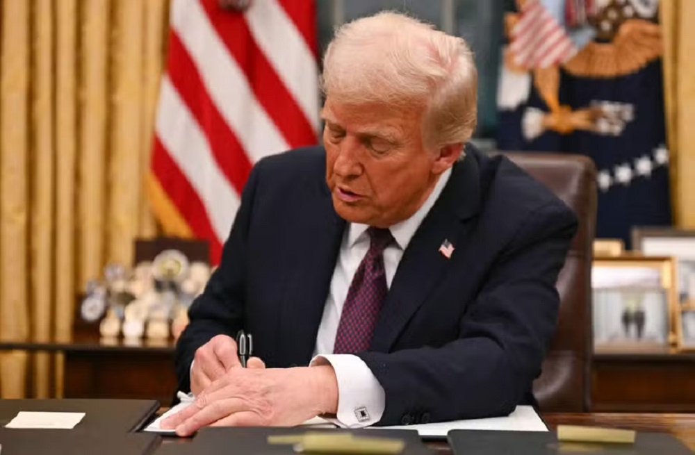 presidente dos Estados Unidos, Donald Trump, assinando papel, de terno e gravata, com bandeira dos EUA ao fundo