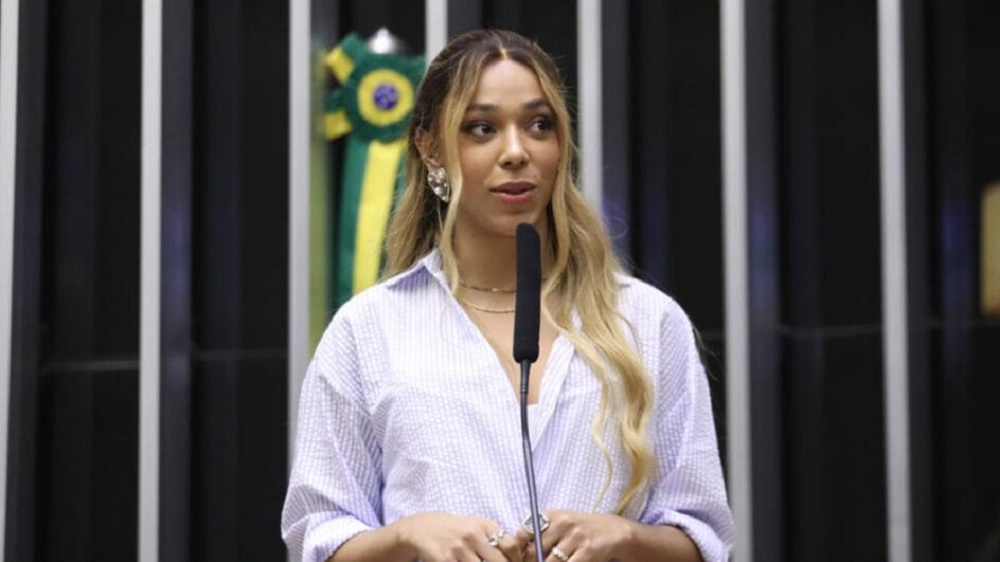 Erika Hilton (PSOL-SP) de roupa branca, séria, perto de microfone, na Câmara dos Deputados