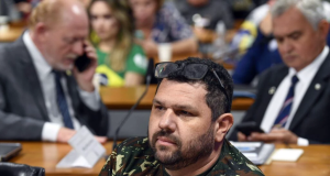 Oswaldo Eustáquio Filho de roupa camuflada, sem olhar pra câmera, sentado, sério, cercado de pessoas que aparecem desfocadas na foto