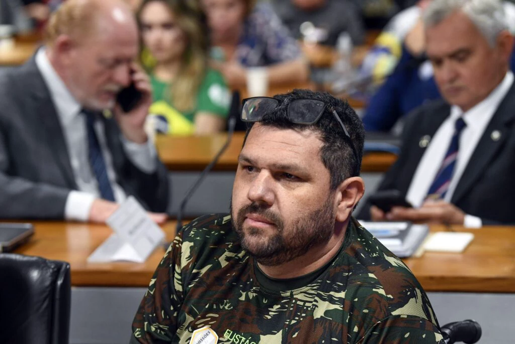 Oswaldo Eustáquio Filho de roupa camuflada, sem olhar pra câmera, sentado, sério, cercado de pessoas que aparecem desfocadas na foto