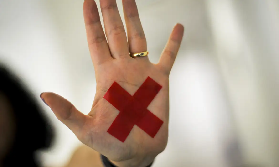 Mão feminina aberta, com aliança dourada e X vermelho na palma da mão