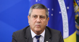general Walter Braga Netto sério, de terno e gravata, com bandeira do Brasil ao fundo, sem olhar para a câmera