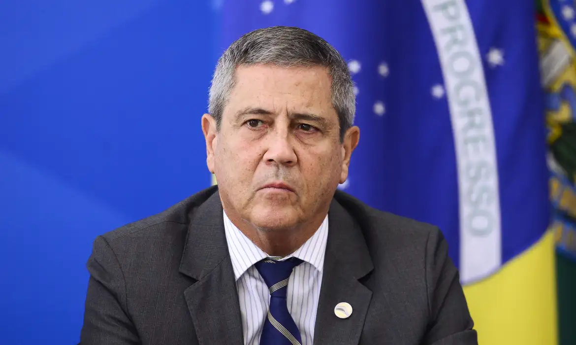 general Walter Braga Netto sério, de terno e gravata, com bandeira do Brasil ao fundo, sem olhar para a câmera