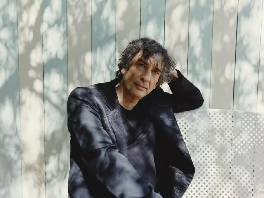 O escritor Neil Gaiman com cabeça apoiada em uma das mãos, de roupa social escura, sério, encostado em parede de madeira