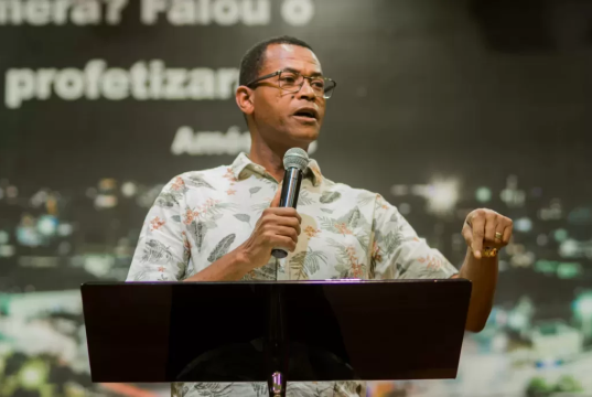 pastor Samuel Bello pregando, segurando microfone, sem olhar para a câmera, sério