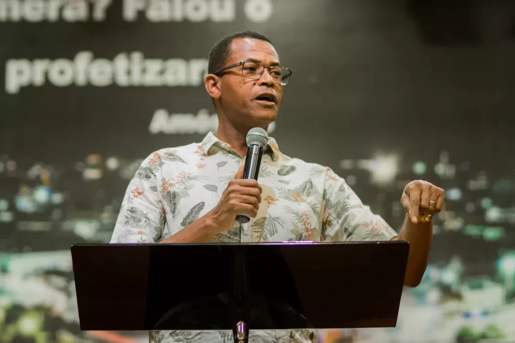 pastor Samuel Bello pregando, segurando microfone, sem olhar para a câmera, sério