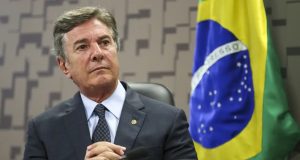 ex-presidente Fernando Collor de Mello sentado perto de bandeira do Brasil, com mãos unidas, sério, olhando para o lado
