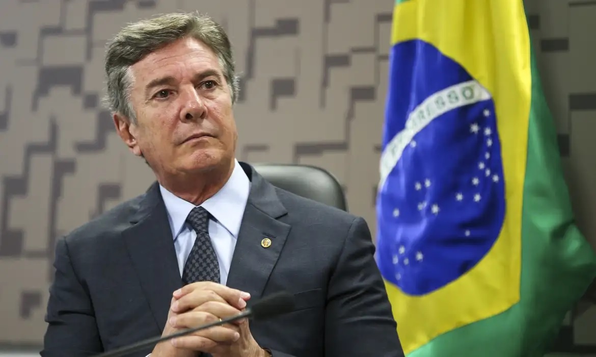 ex-presidente Fernando Collor de Mello sentado perto de bandeira do Brasil, com mãos unidas, sério, olhando para o lado