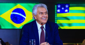 O governador de Goiás, Ronaldo Caiado (União Brasil), sentado, com bandeiras do Brasil e do Goiás ao fundo, olhando para o lado, sorrindo