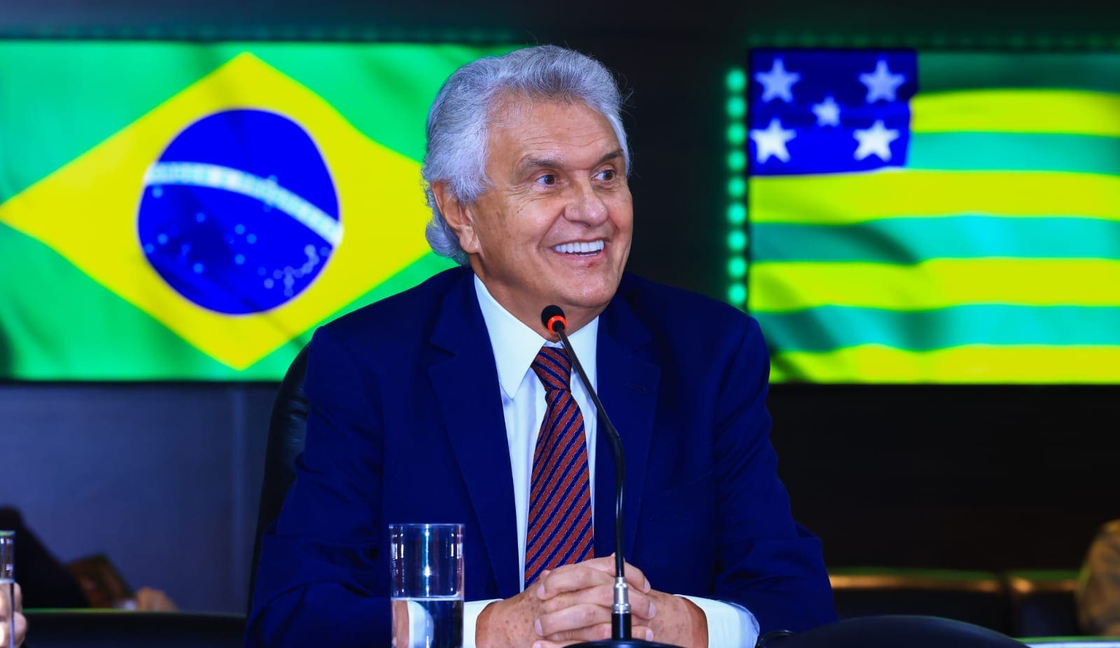 O governador de Goiás, Ronaldo Caiado (União Brasil), sentado, com bandeiras do Brasil e do Goiás ao fundo, olhando para o lado, sorrindo