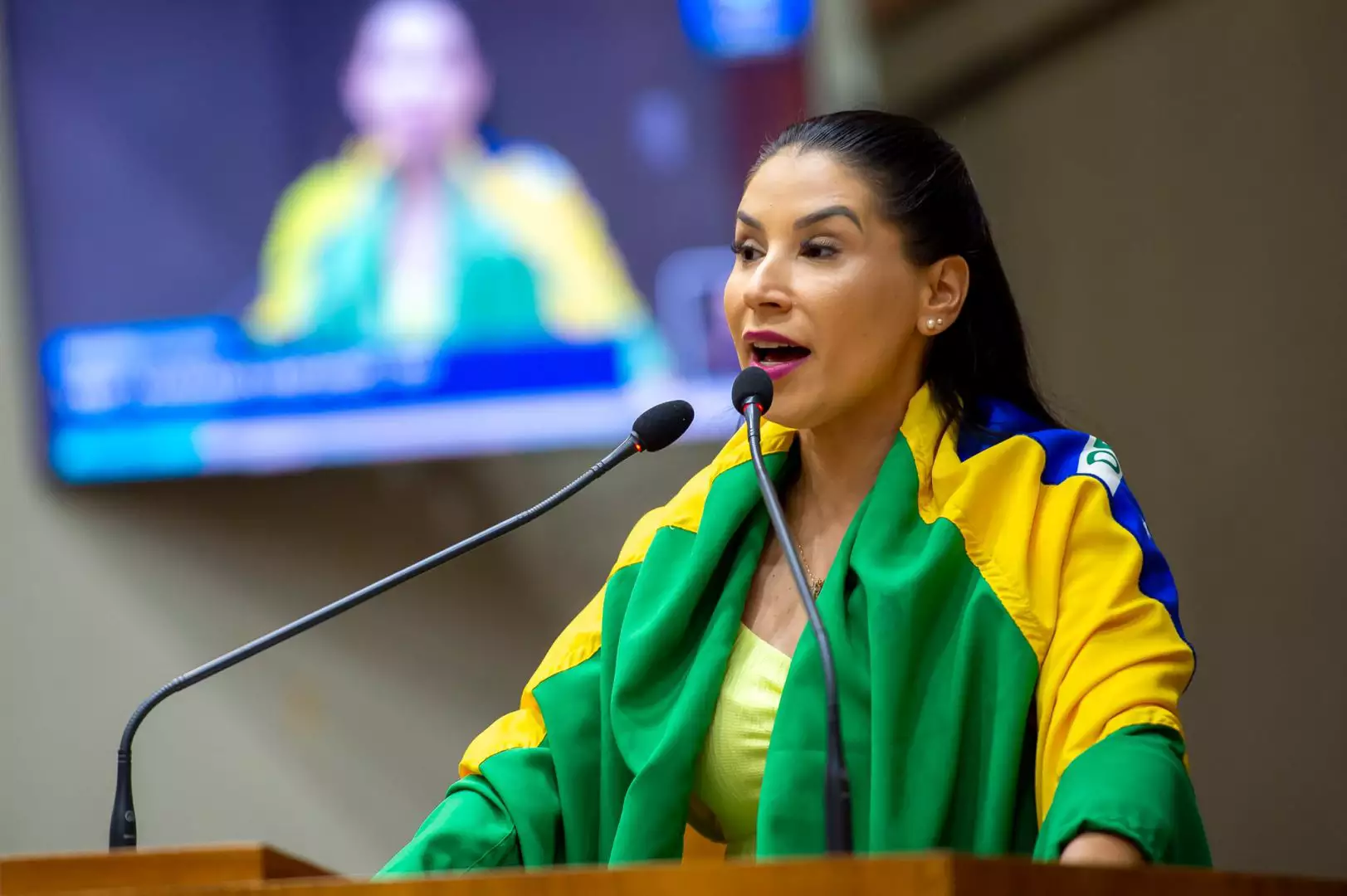 A vereadora Mariana Lescano (Progressistas) de rabo de cavalo, falando em microfone, enrolada em bandeira do Brasil, sem olhar para a câmera