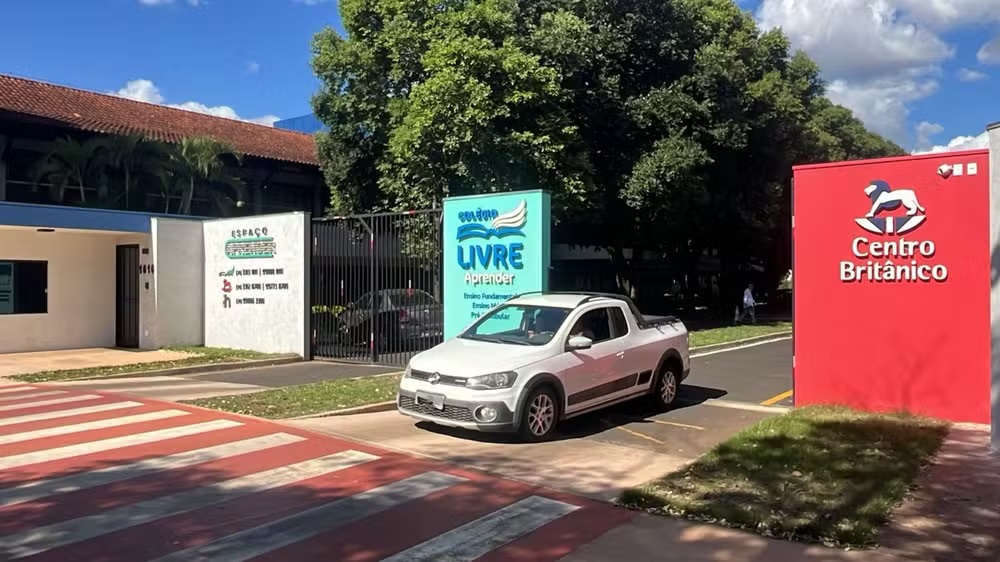 Entrada do Colégio Livre Aprender em dia ensolarado, com carro branco saindo da escola