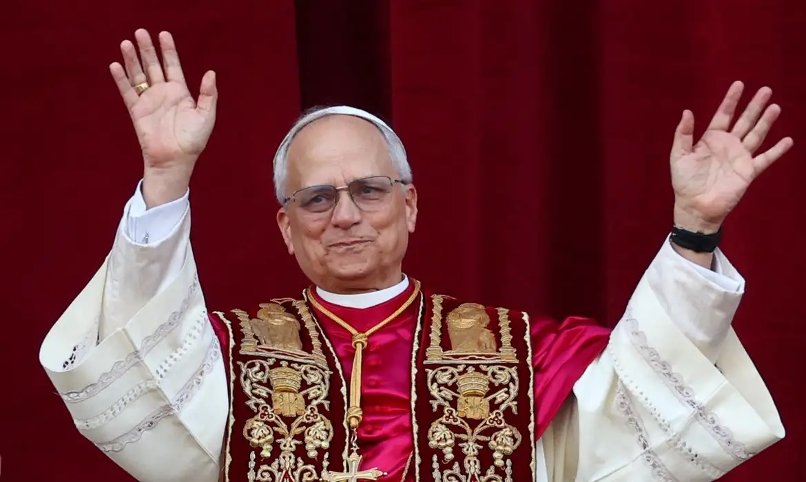 Papa Leão XIV acenando, com sorriso tímido, sem olhar para a câmera
