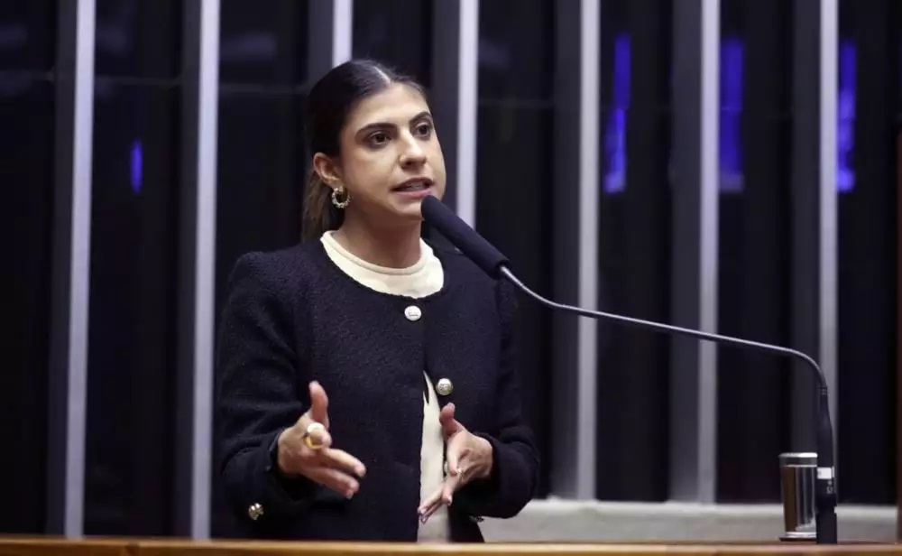 A deputada federal Camila Jara (PT-MS) de roupa preta e branca e rabo de cavalo, falando em microfone e gesticulando