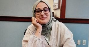 estudante Rumeysa Ozturk de óculos, com rosto apoiado em uma das mãos, sentada perto de mesa, sorrindo de maneira tímida