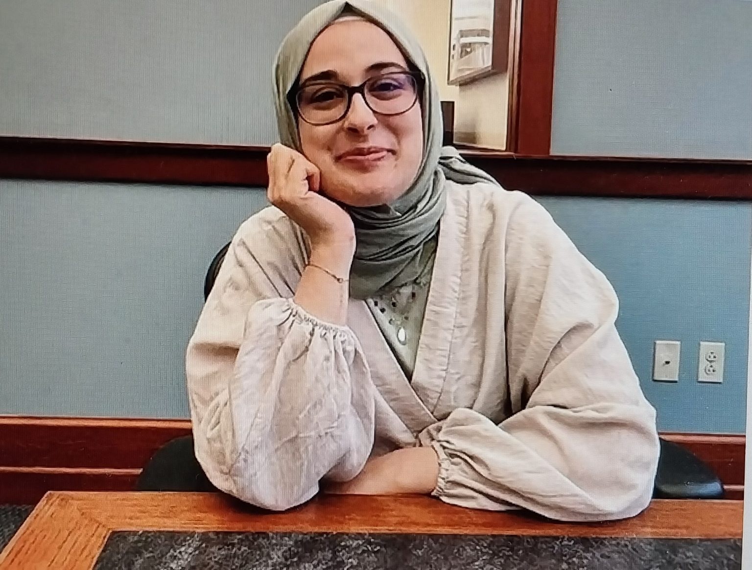 estudante Rumeysa Ozturk de óculos, com rosto apoiado em uma das mãos, sentada perto de mesa, sorrindo de maneira tímida