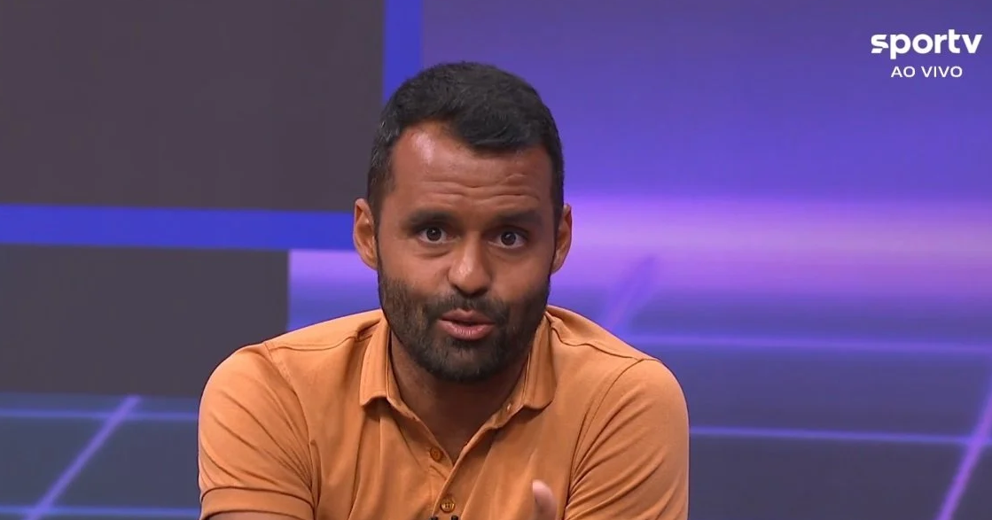 jornalista Conrado Santana falando para a câmera, de roupa coral, sério
