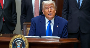 Donald Trump sentado, falando, olhando pra frente, sério
