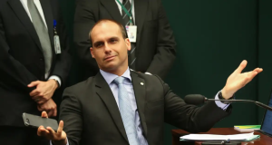 O deputado federal licenciado Eduardo Bolsonaro (PL-SP) sentado, com cara de deboche e os braços abertos, segurando telefone celular