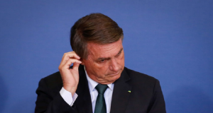 Jair Bolsonaro de cabeça baixa, sério, mexendo na orelha direita