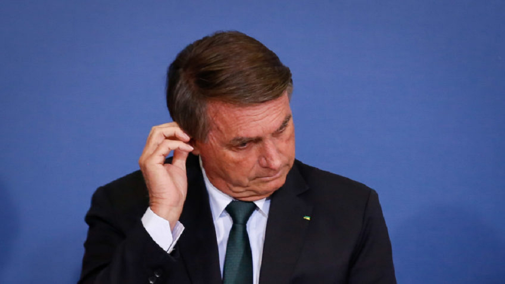 Jair Bolsonaro de cabeça baixa, sério, mexendo na orelha direita