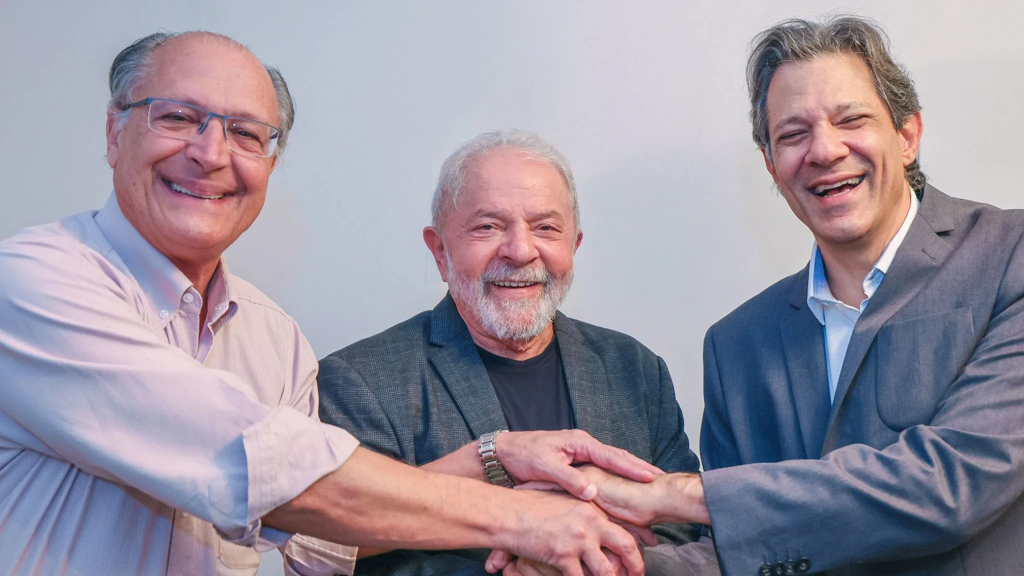 Geraldo Alckmin, Lula e Fernando Haddad sorrindo, em aperto de mãos, em pé, lado a lado