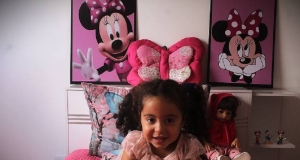 Emanuelly Borges Santos sorrindo, sentada em cama com objetos cor de rosa