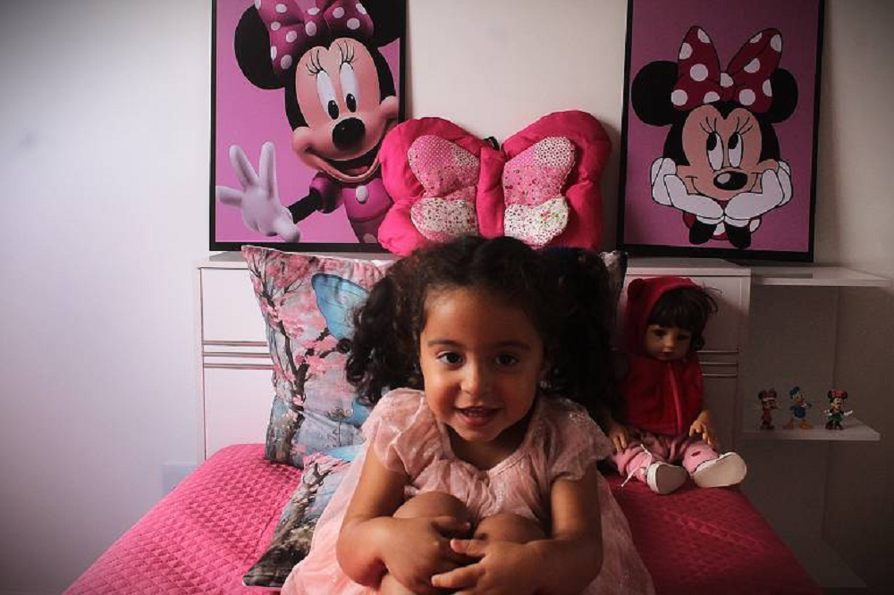 Emanuelly Borges Santos sorrindo, sentada em cama com objetos cor de rosa
