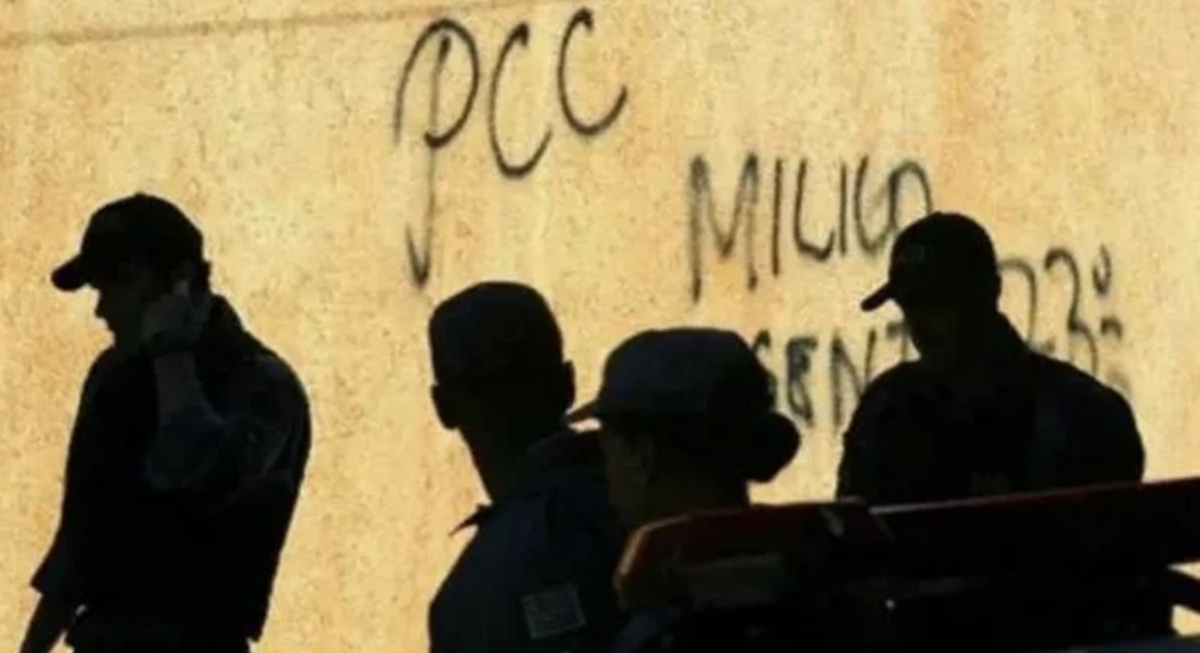 Sombras de quatro pessoas na frente de muro pixado com a sigla do PCC