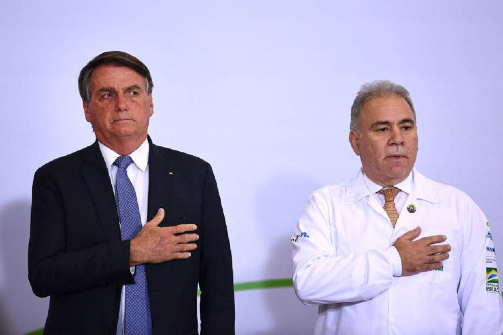 Jair Bolsonaro e Marcelo Queiroga em pé lado a lado, com a mão no peito, sérios