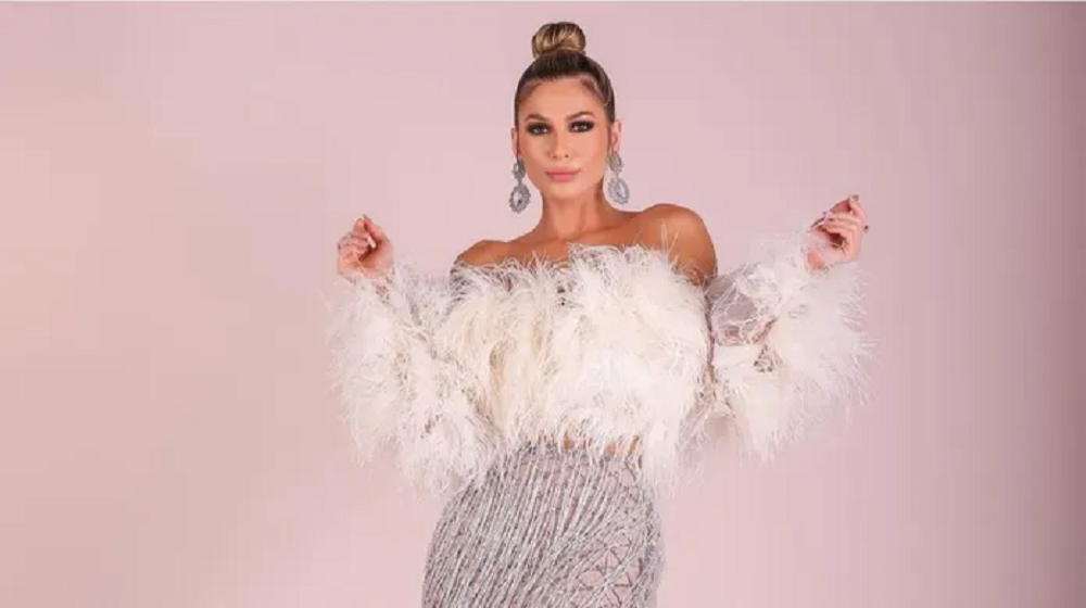 Lívia Andrade séria, de coque, posando com roupa branca com plumas e paetês, em pé, olhando para a câmera