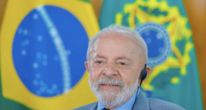 Lula sorrindo, em close, sem olhar para a câmera, com bandeiras ao fundo