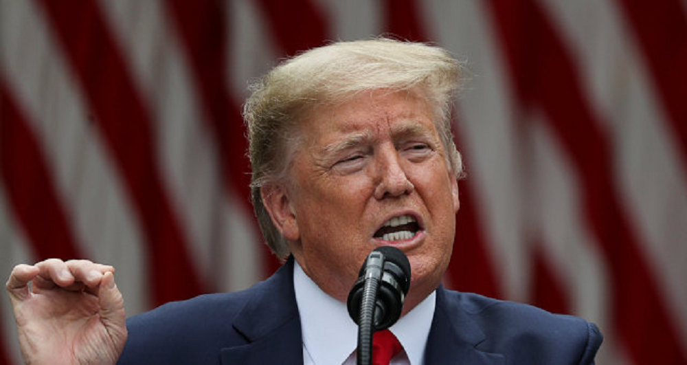 Donald Trump, presidente dos Estados Unidos, falando em microfone com cara de raiva, gesticulando, sem olhar para a câmera, com bandeira dos EUA desfocada ao fundo
