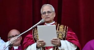 Papa Leão XIV falando em microfone, sério, olhando pra frente, em pé, segurando papel
