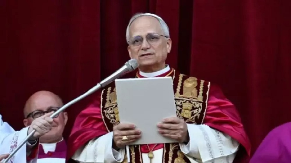 Papa Leão XIV falando em microfone, sério, olhando pra frente, em pé, segurando papel