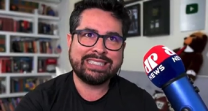 O ex-apresentador da Jovem Pan Paulo Figueiredo fazendo careta, de roupa preta e óculos, perto de microfone da Jovem Pan News