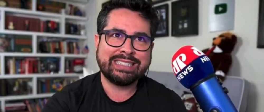 O ex-apresentador da Jovem Pan Paulo Figueiredo fazendo careta, de roupa preta e óculos, perto de microfone da Jovem Pan News