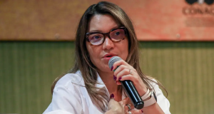 primeira-dama Janja da Silva de roupa branca, óculos de grau e cabelo solto, falando em microfone, séria, com a mão no peito