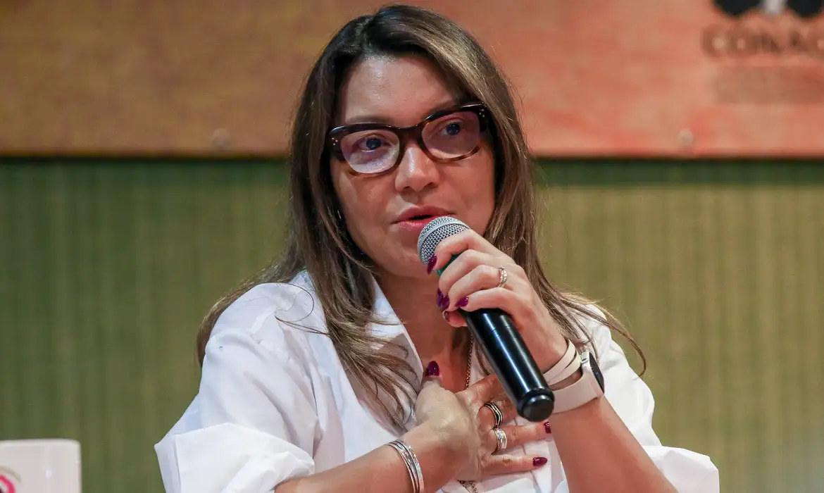 primeira-dama Janja da Silva de roupa branca, óculos de grau e cabelo solto, falando em microfone, séria, com a mão no peito