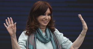 Cristina Kirchner sorrindo, de cabelo solto, com blusa de manga e cachecol, acenando e sorrindo, sem olhar para a câmera