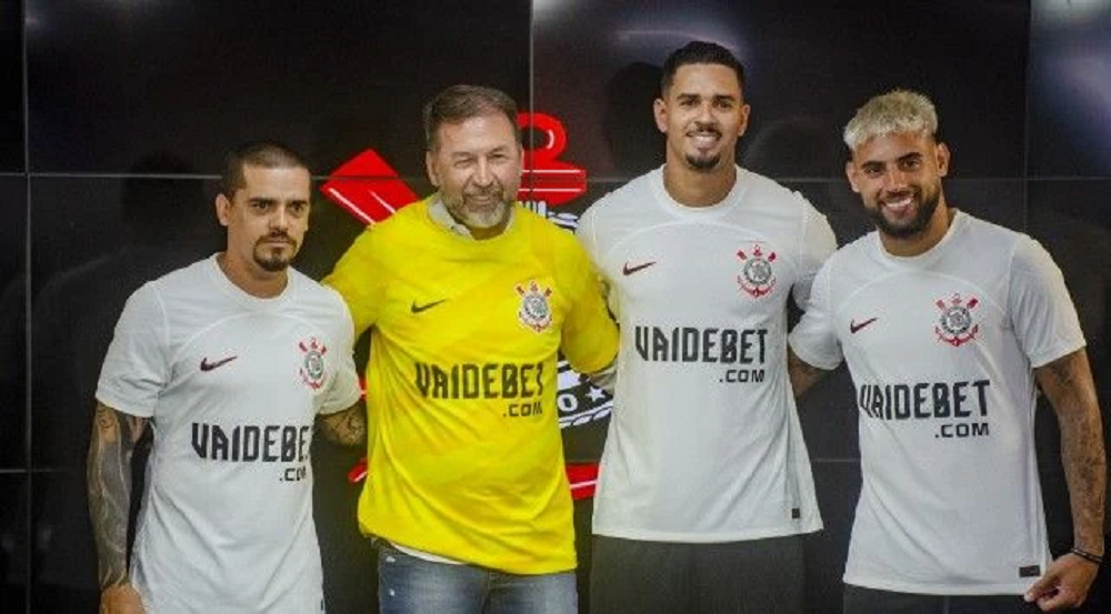 Quatro homens lado a lado com camisetas do Corinthians com anúncio da Vai de Bet