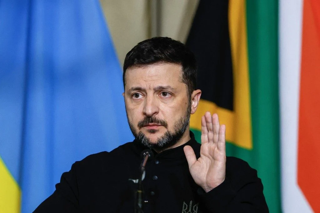 Volodymyr Zelensky de roupa preta, sério, com a mão esquerda levantada, sem olhar para a câmera