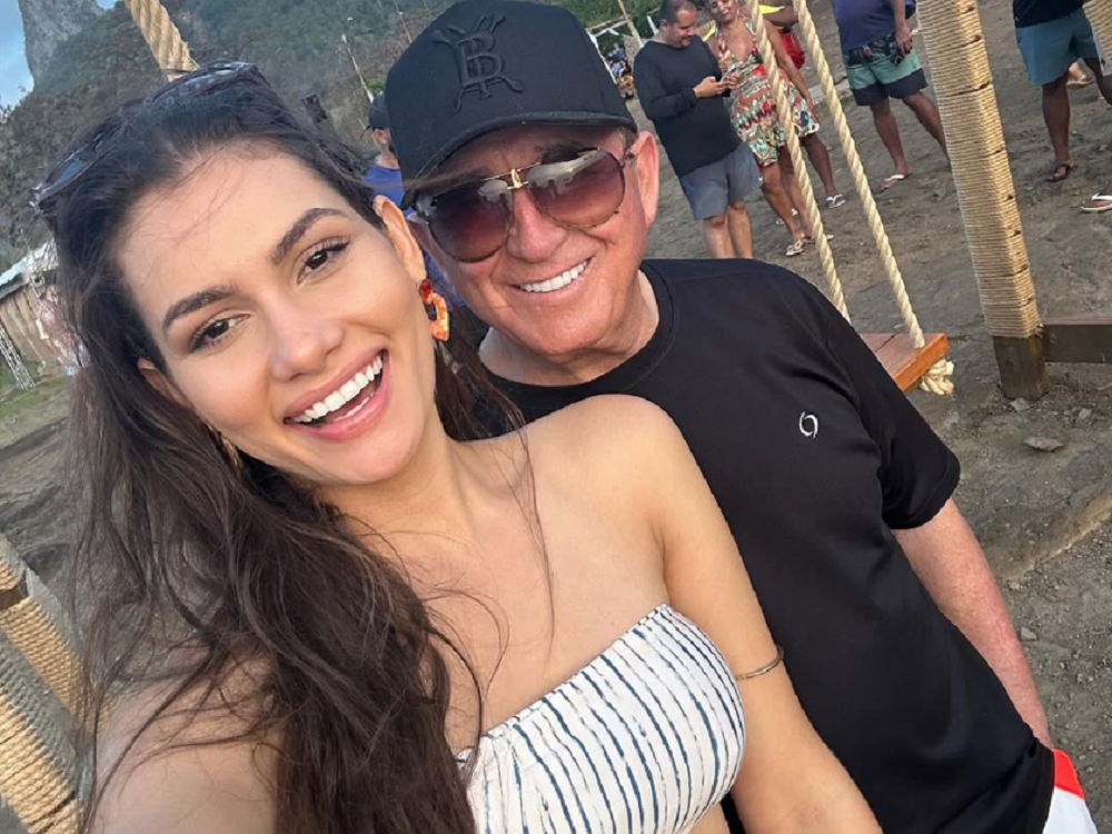 O cantor Amado Batista e a modelo Calita Franciele sorrindo para foto, ele de boné e óculos escuros